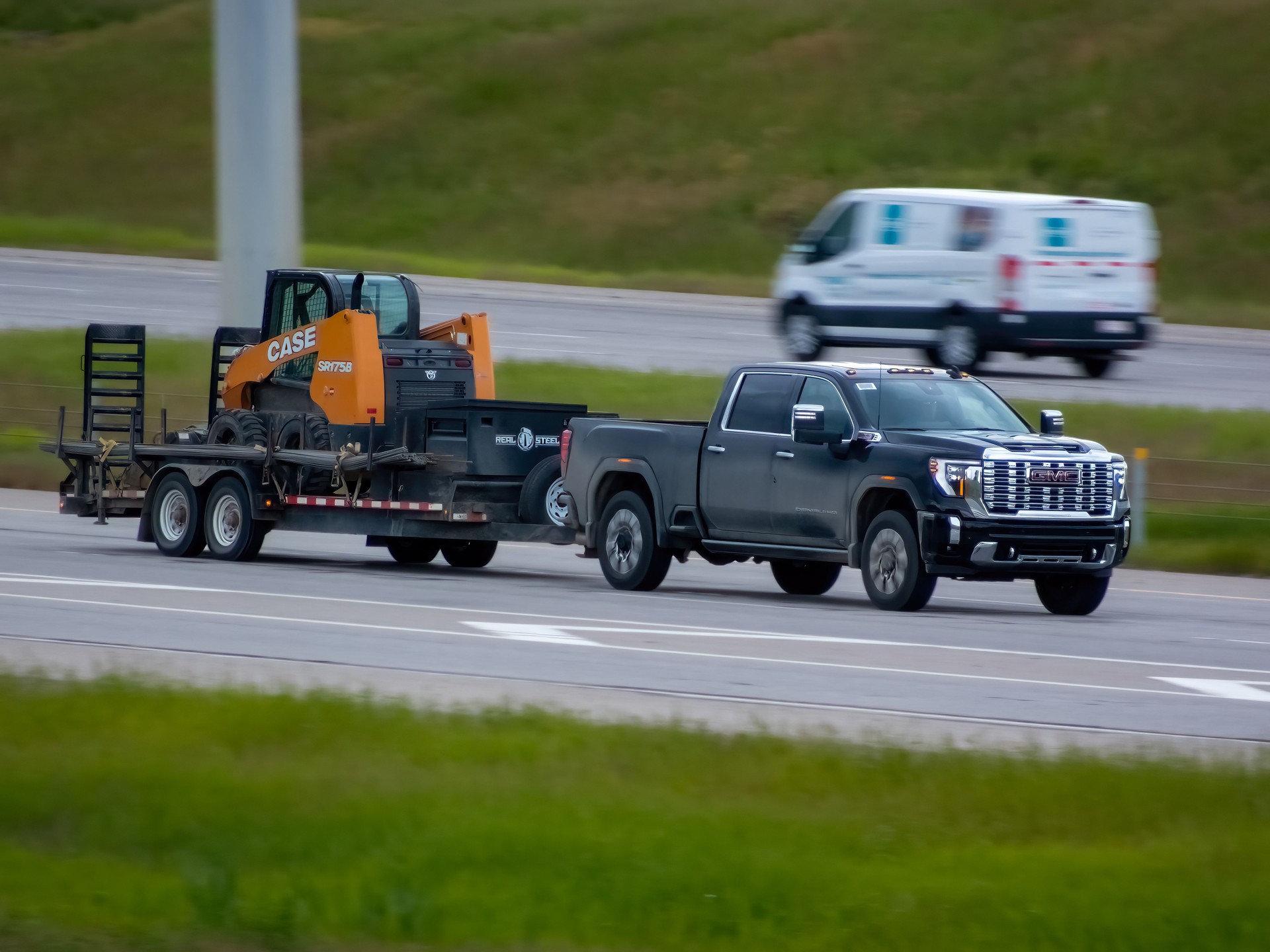 A black GMC Sierra pickup truck tows a black trailer carrying a construction backhoe machine on a highway.